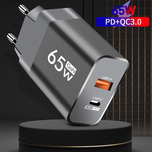 NomadDesk 65W GaN ultra-compact charger in matte charcoal grey with foldable prongs plugged into a minimalist wall next to a portable monitor setup.